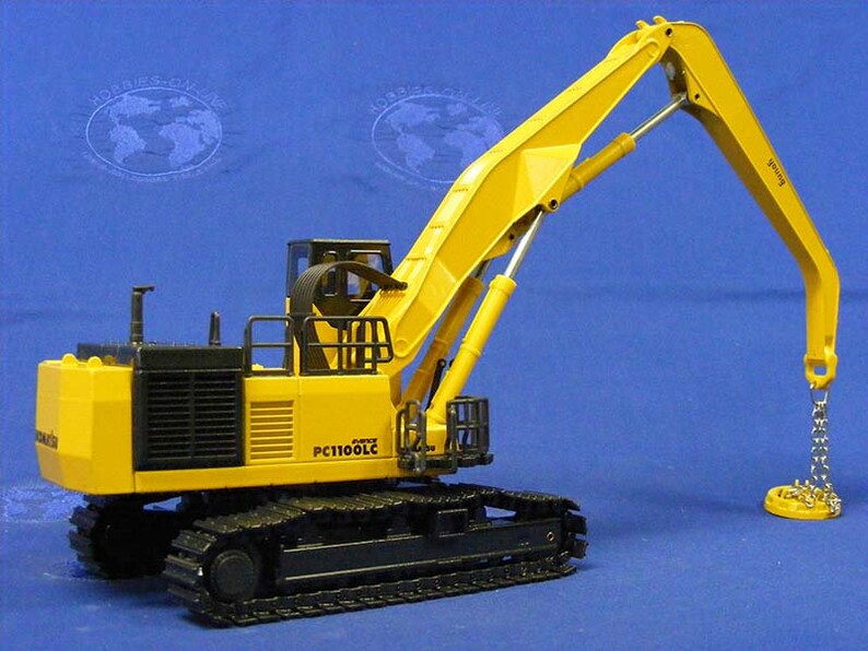 May include: Yellow and black toy model of a Komatsu PC1100LC excavator with a magnetic grapple. The excavator has a long yellow arm with a black chain and a round magnetic plate at the end.