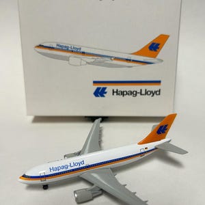May include: A white, orange, and blue Airbus A310-300 airplane model with the Hapag-Lloyd logo. The model is made of metal and is in a 1:500 scale.