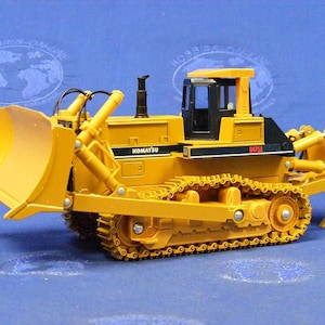 May include: Yellow and black toy bulldozer with a large blade and a Komatsu D75A-1 label. The bulldozer is on a blue surface.