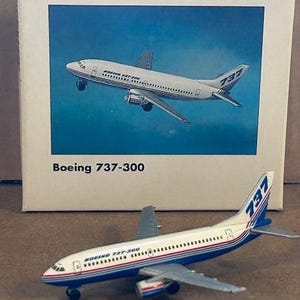 May include: A die-cast Boeing 737-300 model airplane with a white fuselage, blue and red stripes, and 737 branding on the tail. It is displayed in front of its original white cardboard box featuring the text BOEING FLEET and Boeing 737-300.