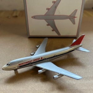 Könnte beinhalten: Ein silbernes und rotes Spielzeugflugzeug mit dem Text "NORTHWEST ORIENT" auf der Seite. Das Modellflugzeug hat vier Triebwerke und eine rote Heckflosse. Im Hintergrund befindet sich eine Schachtel mit einer Zeichnung des Flugzeugs. Auf der Schachtel steht die Nummer "747".