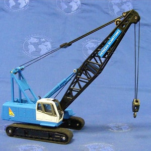 Op de afbeelding: Een blauwe en zwarte speelgoedkraan met de tekst "HITACHI SUMITOMO" op de arm. De kraan staat op een blauwe ondergrond.