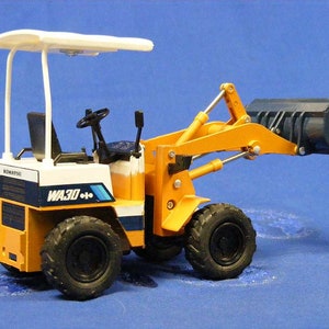 May include: A yellow and white Komatsu WA30-6 wheel loader toy with a black bucket. The toy has a white roll cage and a black steering wheel.