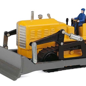 May include: A yellow toy bulldozer with a gray blade and black tracks. A small figure in blue clothing is seated in the driver's seat. The toy has a detailed design, including a radiator grill and exhaust pipe.