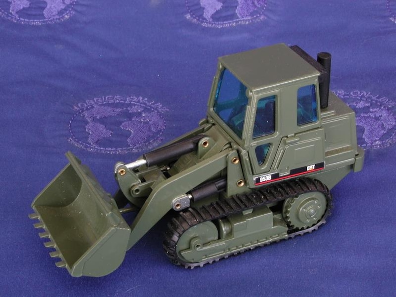 K&ouml;nnte beinhalten: Olivgr&uuml;ner Spielzeugbagger mit Frontschaufel und schwarzen Ketten. Die Kabine hat blau get&ouml;nte Scheiben und das Wort "CAT" ist auf der Seite sichtbar. Die Modellnummer "953B" ist ebenfalls sichtbar.