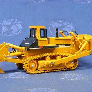 May include: A yellow Komatsu D65A bulldozer toy with a black cab and a large yellow blade. The bulldozer is on a blue surface.
