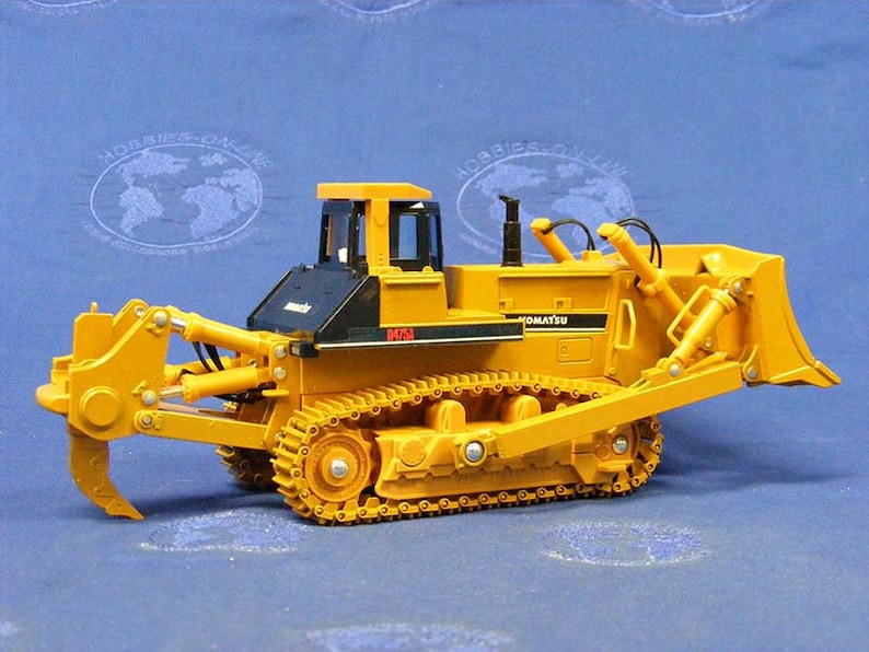 May include: A yellow Komatsu D65A bulldozer toy with a black cab and a large yellow blade. The bulldozer is on a blue surface.