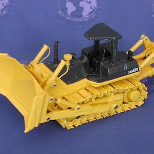 May include: A yellow and black toy bulldozer with a black cab and a yellow blade. The bulldozer has a D155XX label on the side.