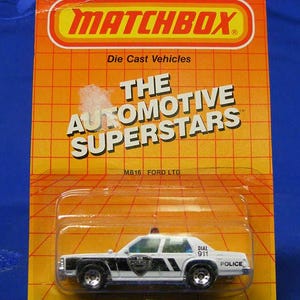 May include: Matchbox die-cast toy car of a Ford LTD police vehicle. The white car has black stripes and a police shield on the doors. The packaging is orange with the words "THE AUTOMOTIVE SUPERSTARS".