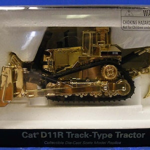 Op de afbeelding: Een goudkleurig die-cast model van een Caterpillar D11R Track-Type Tractor. Het model bevindt zich in een transparante kunststof display case met een witte achtergrond. Het model heeft een zwarte rupsband en een zwarte blad. Het model is gelabeld "Cat D11R Track-Type Tractor" en "Collectible Die-Cast Scale Model Replica".