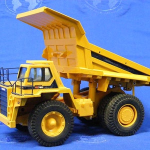 Pu&ograve; includere: Autocarro ribaltabile giocattolo giallo con una gabbia di sicurezza in metallo nero e il testo "KOMATSU HD785" sul lato. Il camion ha una cassa rialzata e grandi pneumatici neri.