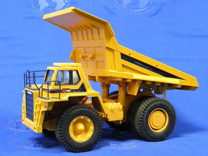 Pu&ograve; includere: Autocarro ribaltabile giocattolo giallo con una gabbia di sicurezza in metallo nero e il testo "KOMATSU HD785" sul lato. Il camion ha una cassa rialzata e grandi pneumatici neri.