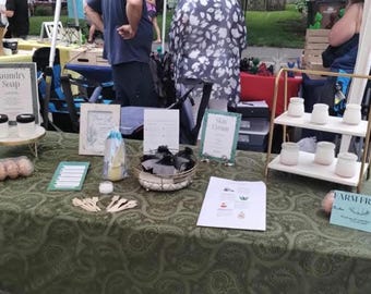 Op de afbeelding: Een groene tafelkleed met verschillende artikelen te koop op een markt. Er zijn potten met wasmiddel, een bord met de tekst "Farm Fresh", een mand met zwarte items en een twee-traps gouden standaard met witte potten.