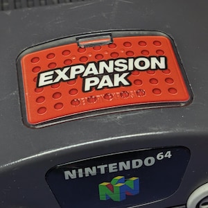 May include: A dark gray Nintendo 64 video game console featuring a red Expansion Pak cover with a perforated pattern and the text EXPANSION PAK in white. The console includes a black NINTENDO 64 logo badge and a gray reset button on the top surface.