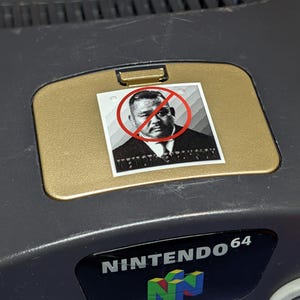May include: A dark gray Nintendo 64 game console featuring a gold-colored expansion pak cover. A square sticker is applied to the cover, displaying a black and white portrait of a person with a red circle and a diagonal line drawn over the image.