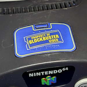 May include: A dark gray Nintendo 64 video game console featuring a blue and yellow sticker on the memory expansion port cover that reads Property of Blockbuster Video. The console includes a power button, a reset button, and a front logo.