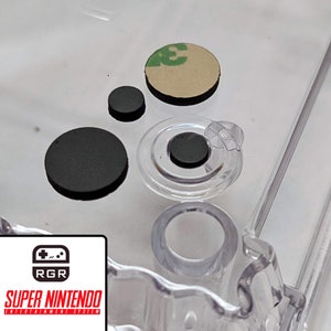 May include: Close-up of a clear plastic Super Nintendo Entertainment System shell with four black rubber buttons and a single beige button with a green sticker. The buttons are used to replace the original buttons on the controller.