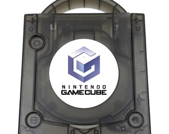 May include: A translucent smoke-gray Nintendo GameCube console featuring a top-loading disc compartment with a circular white logo sticker in the center. The console includes a built-in carrying handle, a power button, a reset button, and an open button.