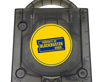 May include: A translucent dark gray video game console with a top-loading disc compartment and a carrying handle. A circular yellow sticker is centered on the lid, featuring a blue ticket graphic with the text PROPERTY OF BLOCKBUSTER VIDEO.