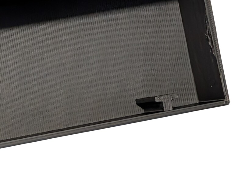 Puede incluir: Primer plano de una superficie gris oscuro texturizada con un patr&oacute;n acanalado. Se ve el borde de un objeto rectangular, con una peque&ntilde;a protuberancia rectangular en el lateral.
