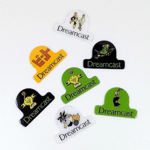 May include: A collection of seven Dreamcast console logo stickers in various colors. The stickers feature the Dreamcast logo and various graphics, including characters and symbols. The stickers are in yellow, black, green, and white.