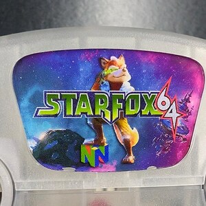 Op de afbeelding: Een grijze Nintendo 64-console met een Star Fox 64-label. Het label toont een cartoon vos-personage tegen een ruimteachtergrond, met de speltitel in groen en geel.