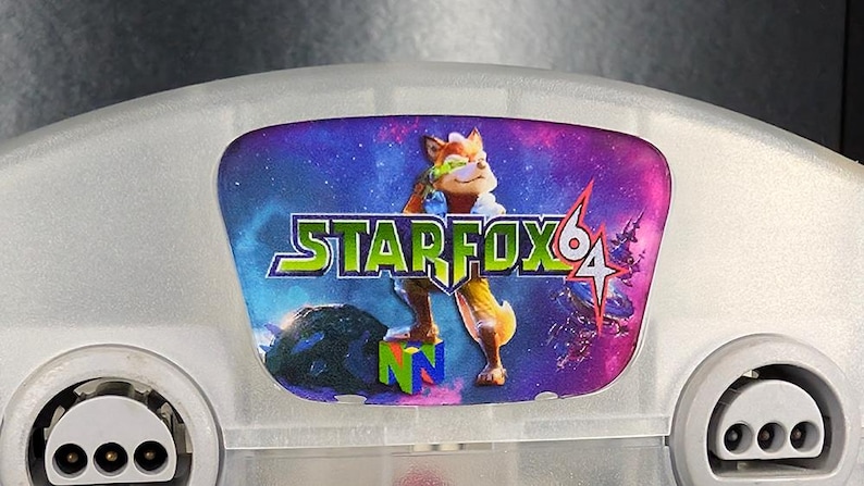 Op de afbeelding: Een grijze Nintendo 64-console met een Star Fox 64-label. Het label toont een cartoon vos-personage tegen een ruimteachtergrond, met de speltitel in groen en geel.