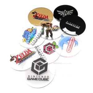 May include: A collection of clear, circular coasters featuring various video game-themed designs. The coasters showcase logos and graphics from games like Zelda, Metroid, and Nintendo GameCube. The coasters are made of clear plastic and are designed to protect surfaces.