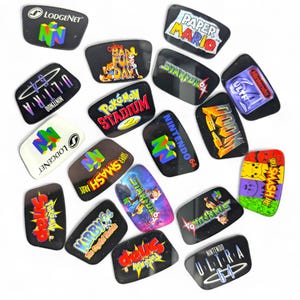 May include: A collection of black, glossy Nintendo 64 game cartridge labels. The labels feature game titles like "Paper Mario," "Star Fox 64," and "Super Smash Bros." in various colors and fonts. The "Nintendo 64" logo is also visible.