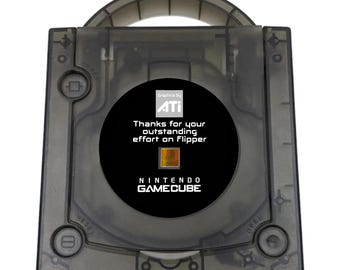 May include: A translucent black Nintendo GameCube console featuring a circular center label. The label displays the ATI logo, the text "Thanks for your outstanding effort on Flipper," a small square graphic, and the Nintendo GameCube brand name.
