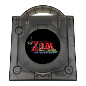 May include: A translucent smoke-gray video game console with a top-loading disc compartment. The center features a circular black emblem with the red and gold text The Legend of Zelda The Wind Waker above stylized blue waves. It has a carry handle.