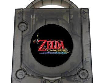 May include: A translucent smoke-gray video game console with a top-loading disc compartment. The center features a circular black emblem with the red and gold text The Legend of Zelda The Wind Waker above stylized blue waves. It has a carry handle.