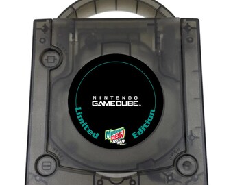 May include: A translucent dark gray Nintendo GameCube console featuring a circular black center disc cover. The disc cover displays the text NINTENDO GAMECUBE, Limited Edition, and a Mountain Dew Baja Blast logo. The console includes a top handle.