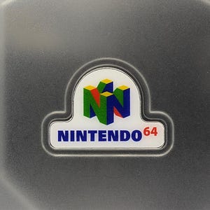May include: A close-up of a gray surface with a Nintendo 64 logo. The logo features a white background with the words "NINTENDO 64" in blue. Above the text are three colorful, stylized 3D blocks in green, yellow, and blue.