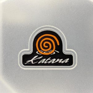 May include: A close-up of a logo with the word "Katana" in white script below a black and orange spiral design. The logo is set against a light gray, slightly textured background.