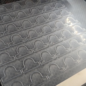 May include: A sheet of clear, transparent plastic with a grid of cut-out shapes resembling the Libra zodiac symbol. The shapes are evenly spaced and arranged in rows and columns. The plastic sheet is on a dark, textured surface.