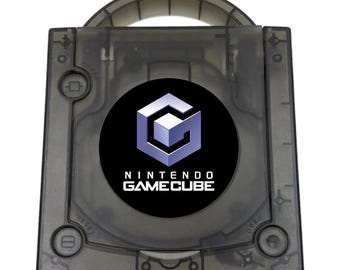 May include: A translucent smoke-gray Nintendo GameCube console featuring a top-loading disc compartment with a circular black logo emblem. The console includes a built-in carrying handle, a power button, a reset button, and an open button.