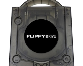 May include: A square, translucent dark gray electronic device housing featuring a circular black center panel with the white text FLIPPY DRIVE printed on it. The unit includes a top handle, a power button, a reset button, and an open button.