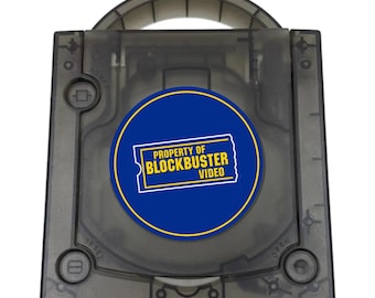 May include: A translucent dark gray game console with a top handle and a circular blue sticker in the center. The sticker features a yellow ticket-shaped graphic with the text PROPERTY OF BLOCKBUSTER VIDEO printed in bold yellow capital letters.