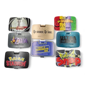 May include: A collection of rectangular, hinged game cartridge cases. Designs include "GoldenEye 007", "Zelda", "Pokemon Stadium", "Pokemon Snap", "Super Smash Bros", "Ultra HDMI", and "Property of Blockbuster Video". One case reads "Ye snooze, Ye lose."