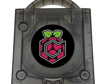 May include: A translucent smoke-gray game console case featuring a circular black emblem in the center. The emblem displays a stylized magenta and black hexagonal pattern topped with two green leaves, outlined in white, resembling a berry.