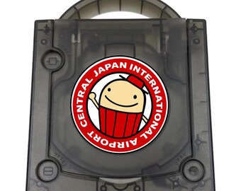 May include: A translucent black game console with a top-loading disc compartment and a carrying handle. A circular sticker is centered on the lid, featuring a cartoon character in a red hat and red clothing with the text CENTRAL JAPAN INTERNATIONAL AIRPORT.