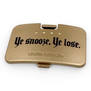 May include: A rectangular gold-colored plastic cover featuring the black printed text "Ye snooze, Ye lose." and the embossed text "MEMORY EXPANSION" below it. The piece includes a small latch mechanism at the top and bottom edge for attachment.