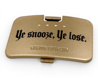 May include: A rectangular gold-colored plastic cover featuring the black printed text "Ye snooze, Ye lose." and the embossed text "MEMORY EXPANSION" below it. The piece includes a small latch mechanism at the top and bottom edge for attachment.