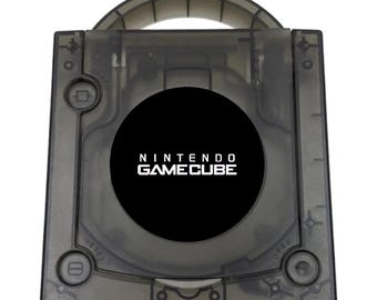 May include: A translucent smoke-gray Nintendo GameCube video game console featuring a top-loading disc compartment with a circular black center logo. The square device includes a power button, a reset button, an open button, and a carrying handle.