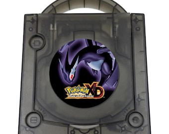 May include: A translucent black GameCube console featuring a circular disc cover graphic of the video game Pokémon XD: Gale of Darkness. The console includes a top handle, a power button, a reset button, and an open button on the front face.