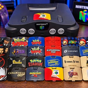 May include: A charcoal gray Nintendo 64 console with a controller and a collection of twenty-four custom jumper pak covers featuring various game titles and logos including Pokemon Snap, Super Mario 64, GoldenEye 007, and Blockbuster Video.