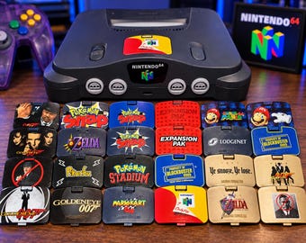 May include: A charcoal gray Nintendo 64 console with a controller and a collection of twenty-four custom jumper pak covers featuring various game titles and logos including Pokemon Snap, Super Mario 64, GoldenEye 007, and Blockbuster Video.