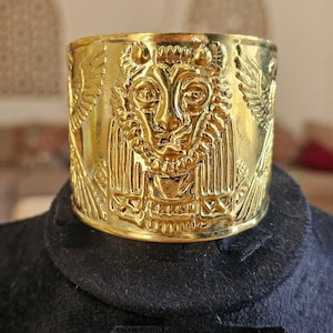 May include: A wide, gold-colored cuff bracelet with an embossed lion's head and winged figures. The cuff is displayed on a black surface, showcasing the intricate details of the jewelry. The design is reminiscent of ancient Egyptian art.