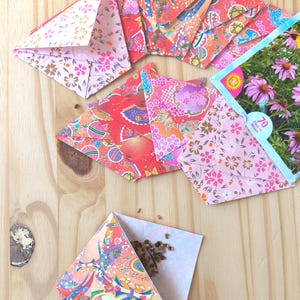 May include: A collection of folded paper seed packets featuring vibrant floral and traditional patterns in shades of pink, orange, and red. One packet is open, revealing small brown seeds, and another shows a picture of purple flowers with the text 70 Days to Bloom.